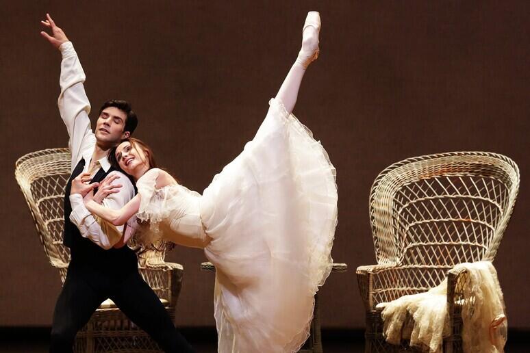 Un dúo de ballet actúa en el escenario; la mujer, con un tutú blanco, extiende con elegancia la pierna, mientras el hombre la sostiene. Al fondo se ven dos sillas de mimbre.