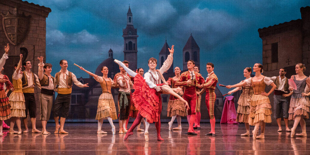 En el escenario, una representación de ballet muestra a bailarines con trajes tradicionales, con una bailarina principal vestida con una falda roja en una pose elegante. El telón de fondo muestra una escena rural bajo un cielo nublado, lo que evoca un ambiente animado y lleno de vida.