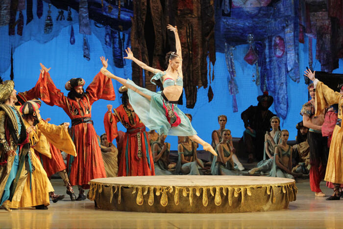 Una bailarina de ballet salta con elegancia sobre el escenario, rodeada de artistas con trajes coloridos y elaborados. El telón de fondo es muy llamativo y crea una atmósfera encantadora.