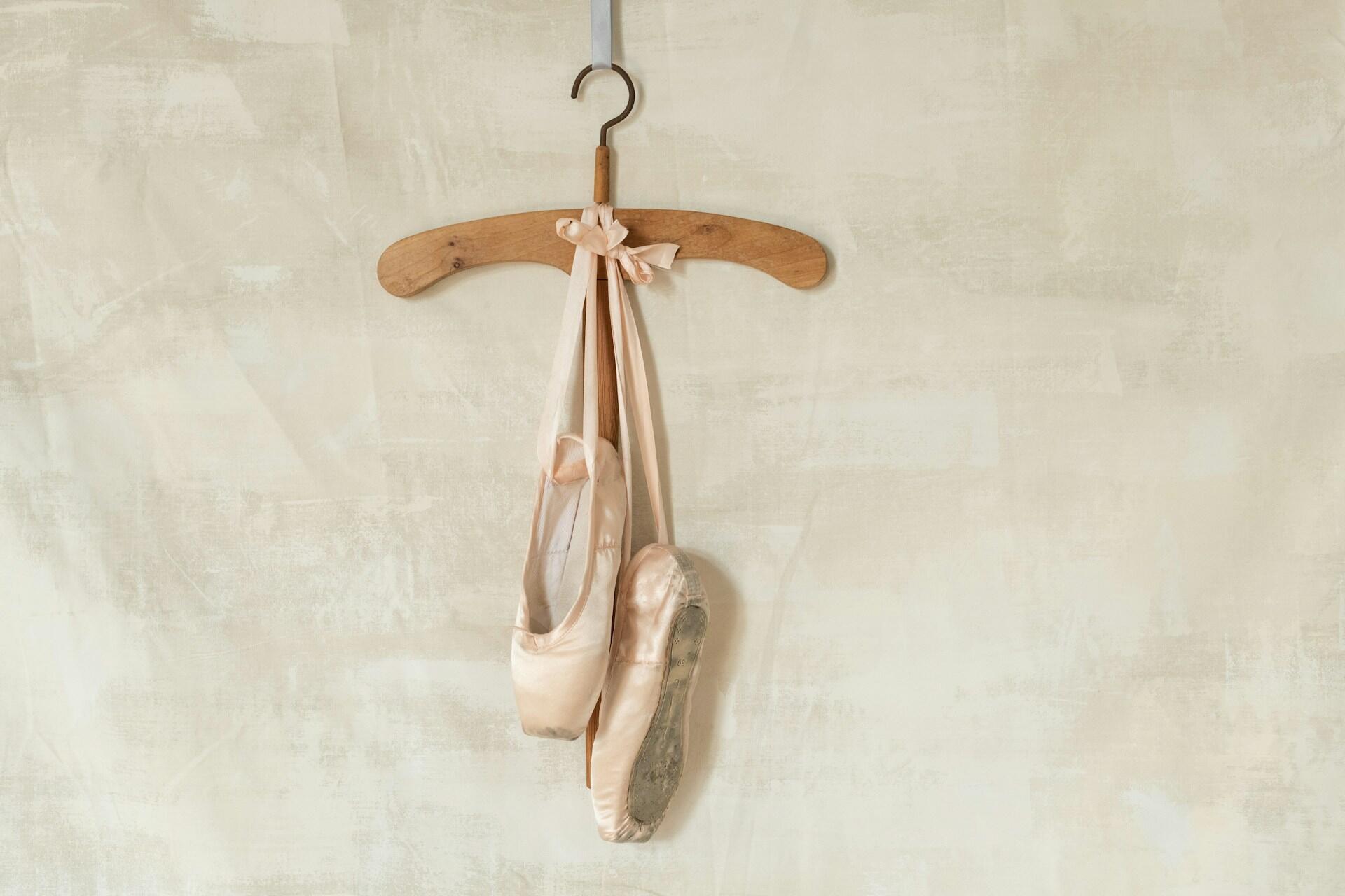 Unas zapatillas de ballet de color rosa pálido cuelgan de una percha de madera sobre un fondo beige claro con textura, lo que evoca una sensación de gracia y elegancia.