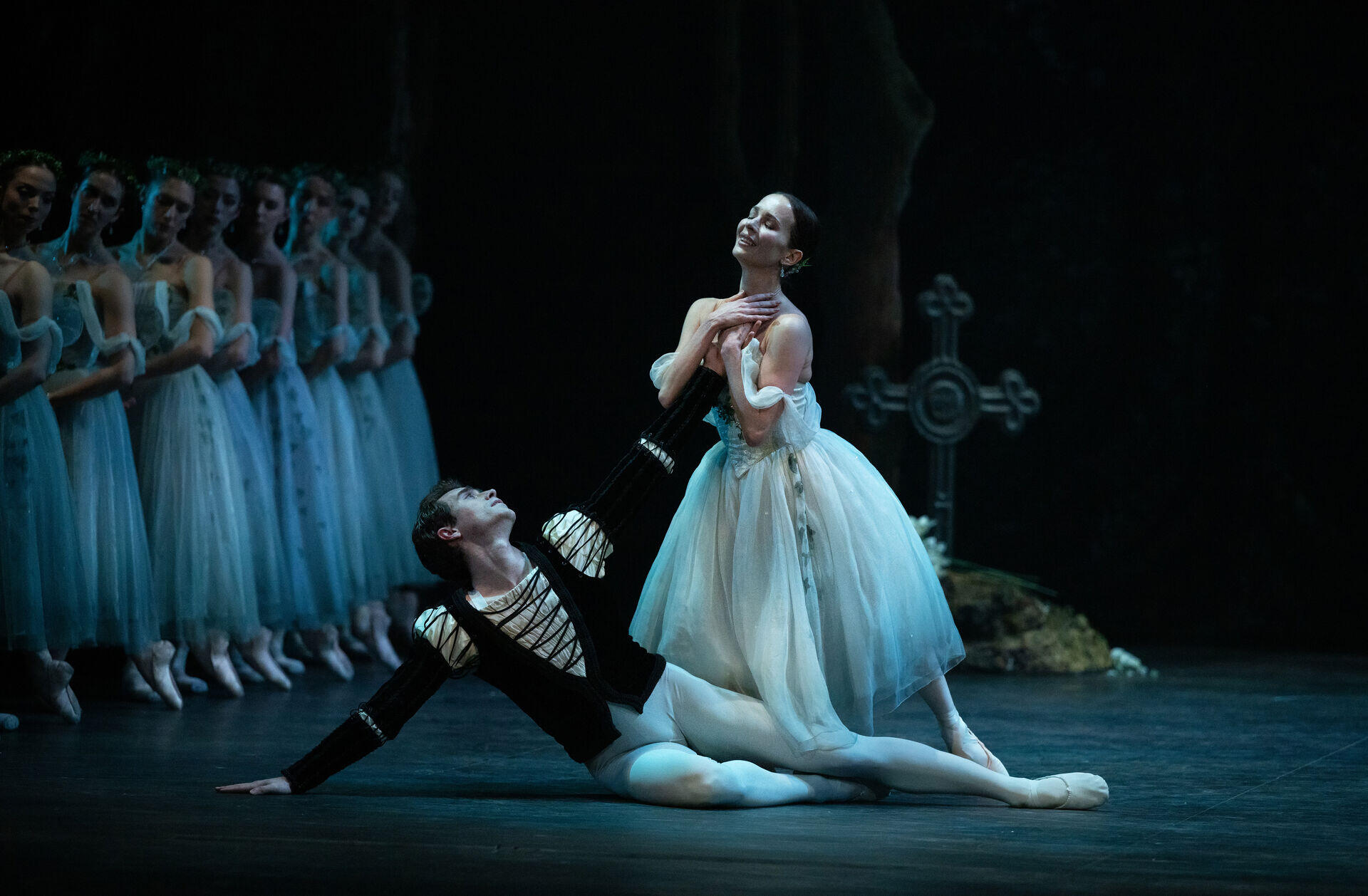 Los bailarines de ballet interpretan una escena emotiva sobre el escenario. Un bailarín se arrodilla y sostiene a una bailarina que permanece de pie con elegancia. Al fondo, una fila de bailarinas con tutús blancos forma parte de un escenario sobrio y evocador.