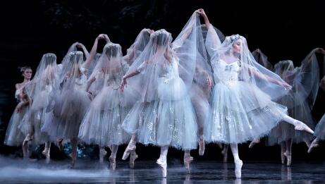 Las bailarinas de ballet, con sus etéreos tutús blancos, actúan con gracia sobre el escenario, creando una escena elegante con velos que ondean al viento y movimientos llenos de aplomo, lo que genera un ambiente de ensueño.