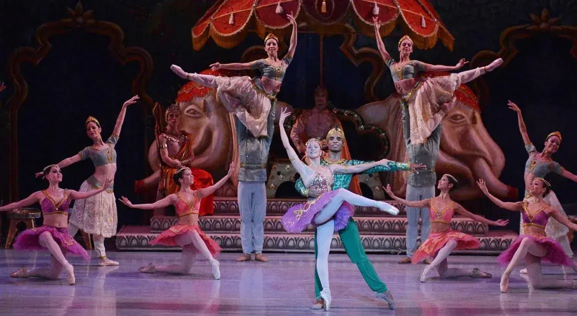 Los bailarines de ballet actúan en el escenario, ataviados con trajes elaborados y coloridos. Dos bailarines realizan una elegante elevación en el centro, rodeados por el resto del grupo. La escena es vibrante y elegante.