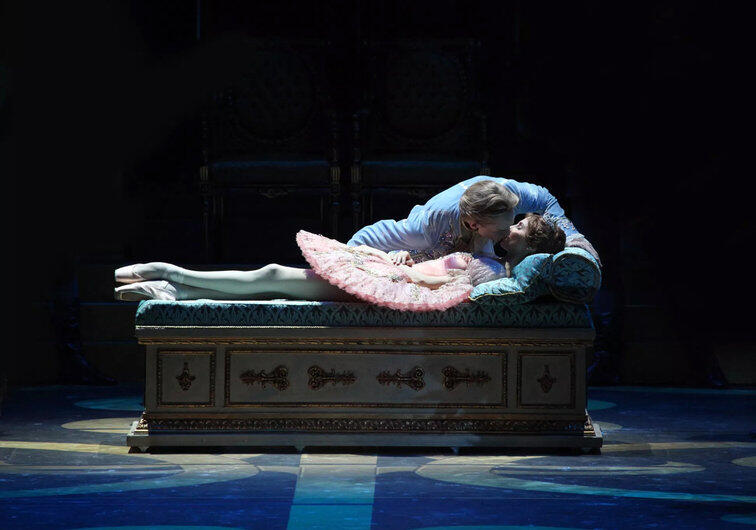 Un príncipe con una túnica azul se inclina sobre una princesa dormida con un tutú rosa en un banco ornamentado, creando una escena de cuento de hadas con un ambiente tierno y mágico.