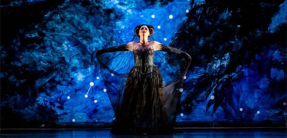 Una artista, vestida con un elegante vestido negro, posa ante un fondo azul intenso salpicado de estrellas. La iluminación dramática y el vestido vaporoso transmiten una sensación de misterio y encanto.