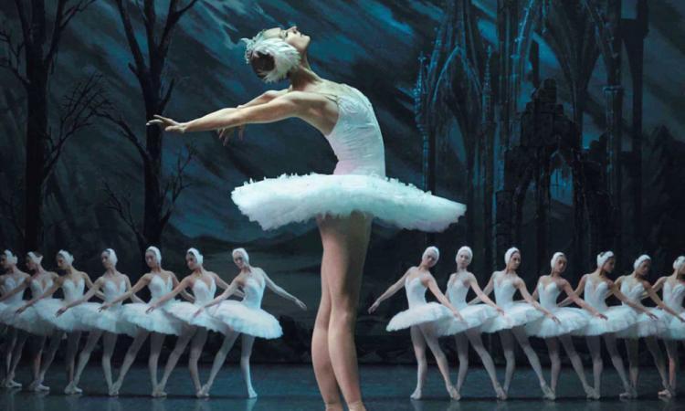 Una bailarina con un tutú blanco actúa en el escenario, adoptando una pose elegante con los brazos extendidos. Detrás de ella, le sigue una fila de bailarinas con un atuendo similar.