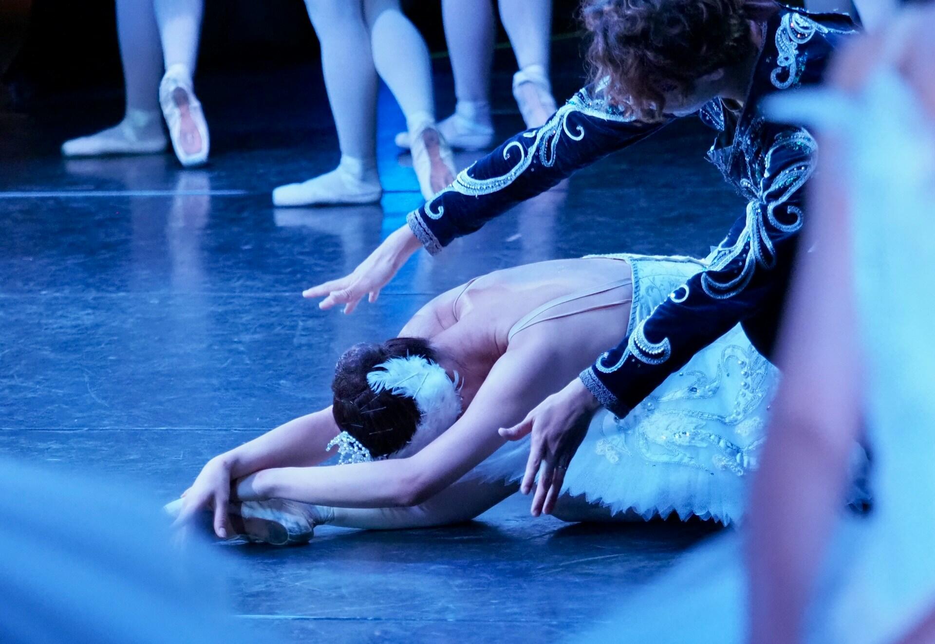 Unos bailarines de ballet, ataviados con elegantes trajes blancos, actúan en un escenario con luz tenue. Una bailarina se estira con gracia en el suelo, mientras otra se mantiene suspendida sobre ella en actitud protectora.