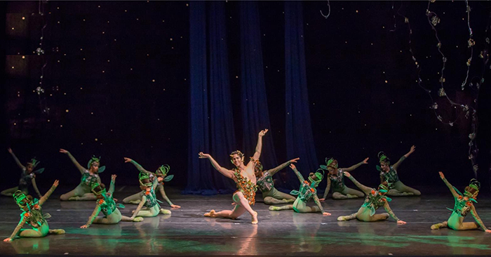 Una bailarina de ballet con un traje colorido posa con elegancia en el centro del escenario, rodeada de bailarines con trajes verdes adornados con hojas, creando una escena forestal llena de fantasía.