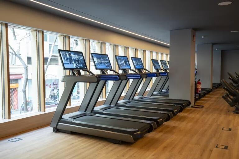 Seis cintas de correr modernas con pantallas digitales en un gimnasio luminoso y espacioso, con suelos de madera y grandes ventanales, que crean un ambiente acogedor y lleno de energía.
