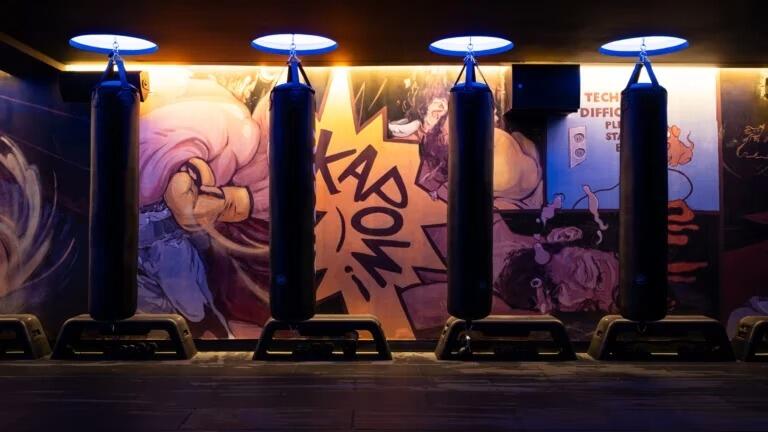 Una sala de gimnasio con cuatro sacos de boxeo alineados frente a un llamativo mural de estilo cómic en el que se representan escenas de acción y la palabra «¡KAPOW!», todo ello bajo una suave iluminación cenital.