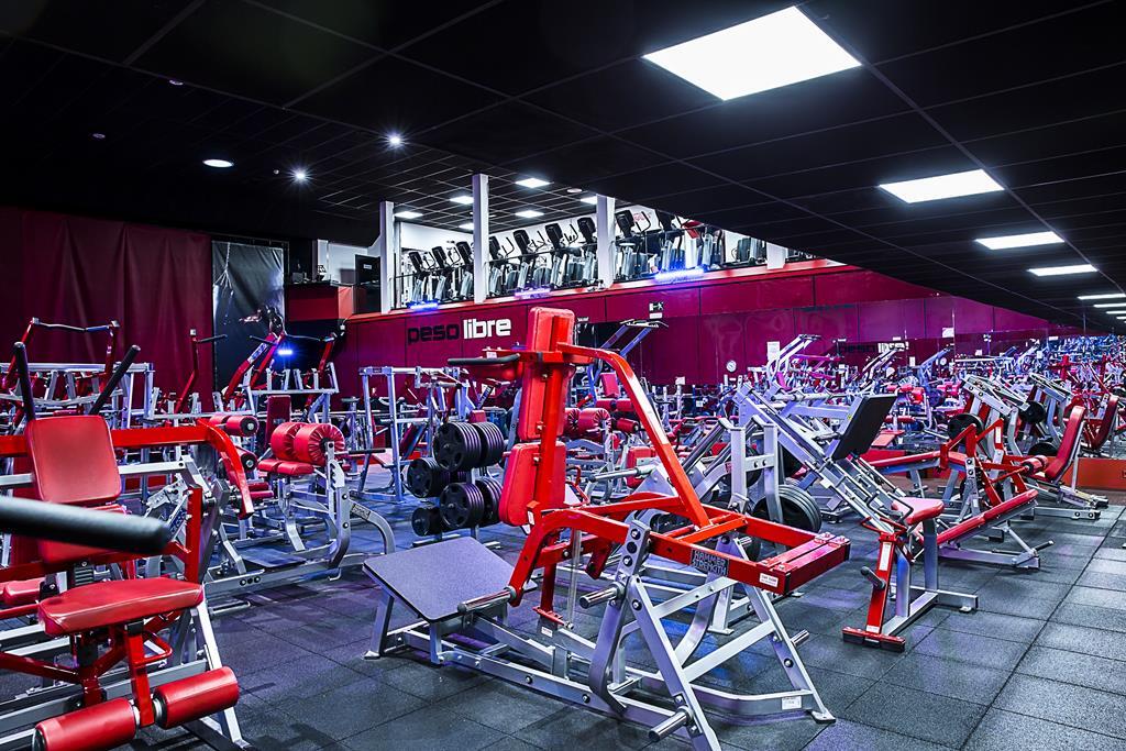Un amplio gimnasio con máquinas de ejercicio rojas y plateadas sobre un suelo negro. Unas luminosas luces de techo iluminan las filas de máquinas, con un balcón en la parte superior.