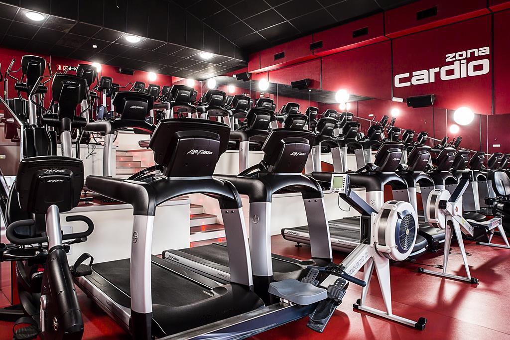 Gimnasio moderno con filas de cintas de correr y bicicletas estáticas negras sobre un suelo rojo. La iluminación brillante y los espejos crean una sensación de energía y amplitud.