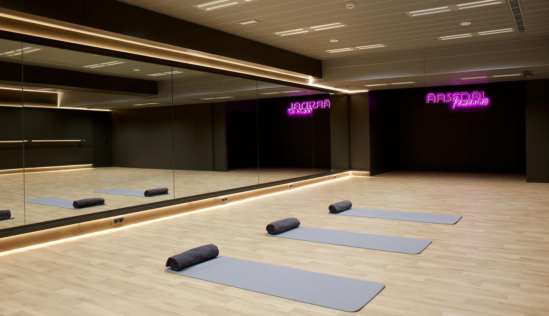 Amplio estudio de yoga con suelos de madera clara, una pared con espejos y tres colchonetas enrolladas. El letrero de neón brilla en color morado, creando un ambiente tranquilo y moderno.