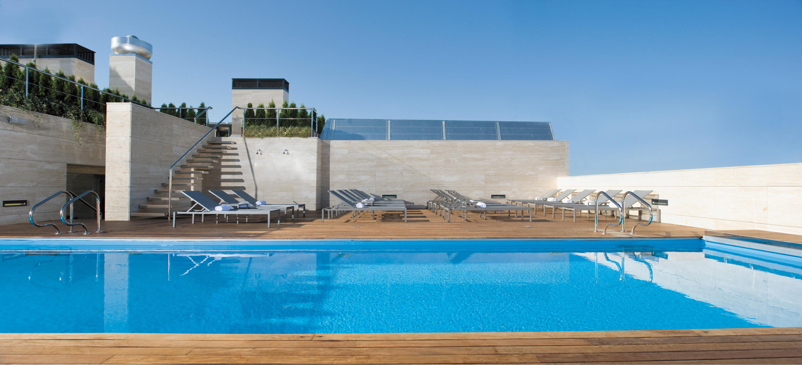 Piscina en la azotea con aguas cristalinas, rodeada de una terraza de madera, con tumbonas vacías y una barandilla de cristal. Un día luminoso y soleado.