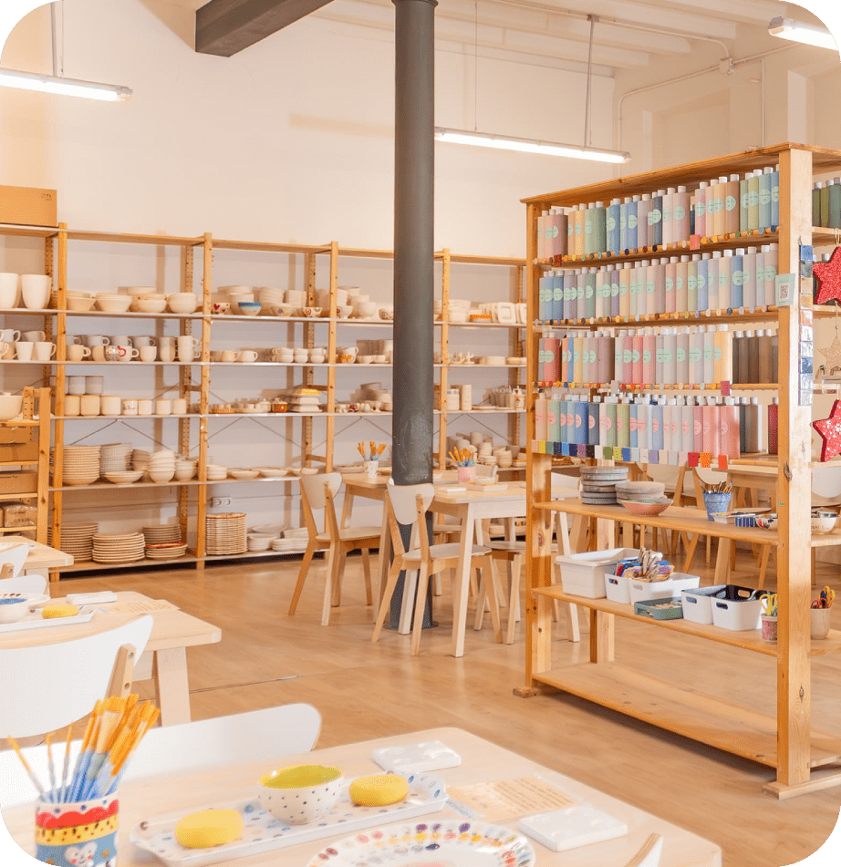 Foto del luminoso taller Ceramicària con estanterías de madera repletas de piezas de cerámica, pinturas de colores y materiales artísticos sobre las mesas para realizar actividades creativas.