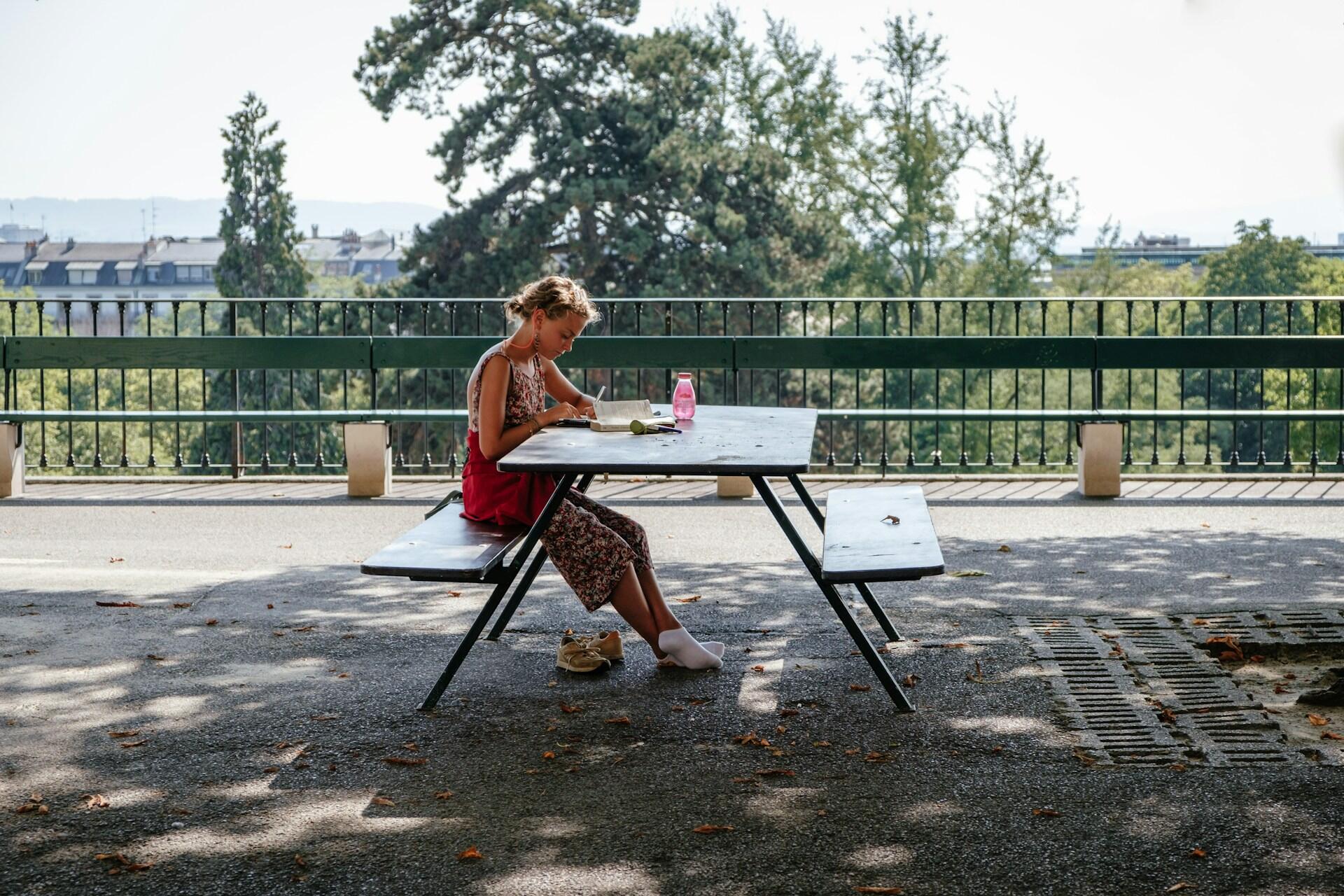 Una mujer está sentada en silencio en una mesa de picnic en un parque, concentrada en escribir. La luz del sol se cuela entre los árboles, proyectando suaves sombras sobre el suelo.