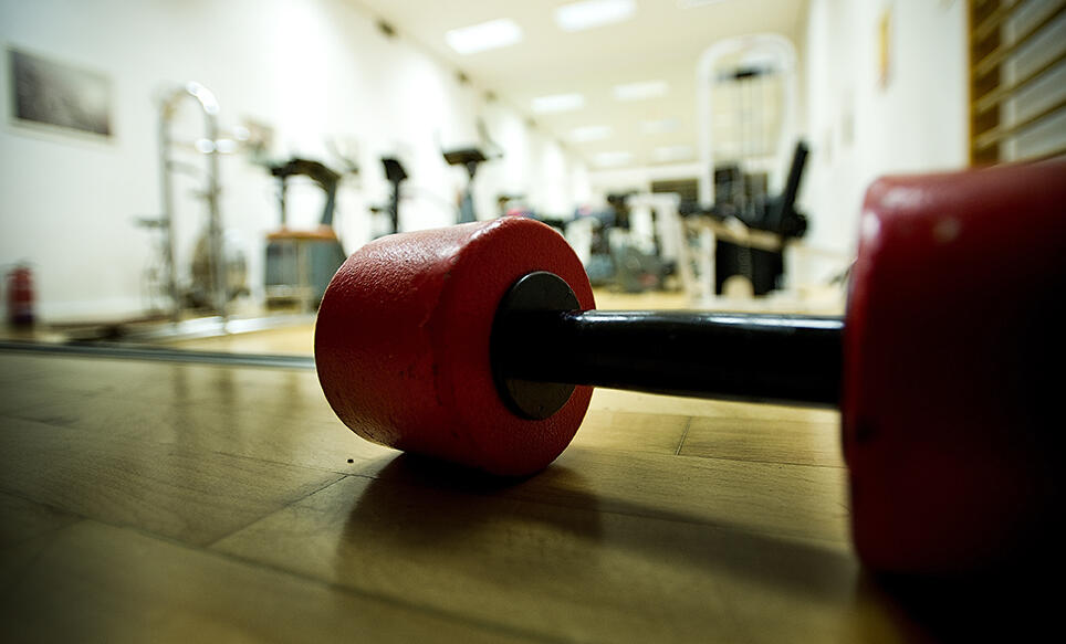 Una mancuerna roja sobre el suelo de madera de un gimnasio, rodeada de máquinas de ejercicio difuminadas, creando un ambiente de concentración y dinamismo.