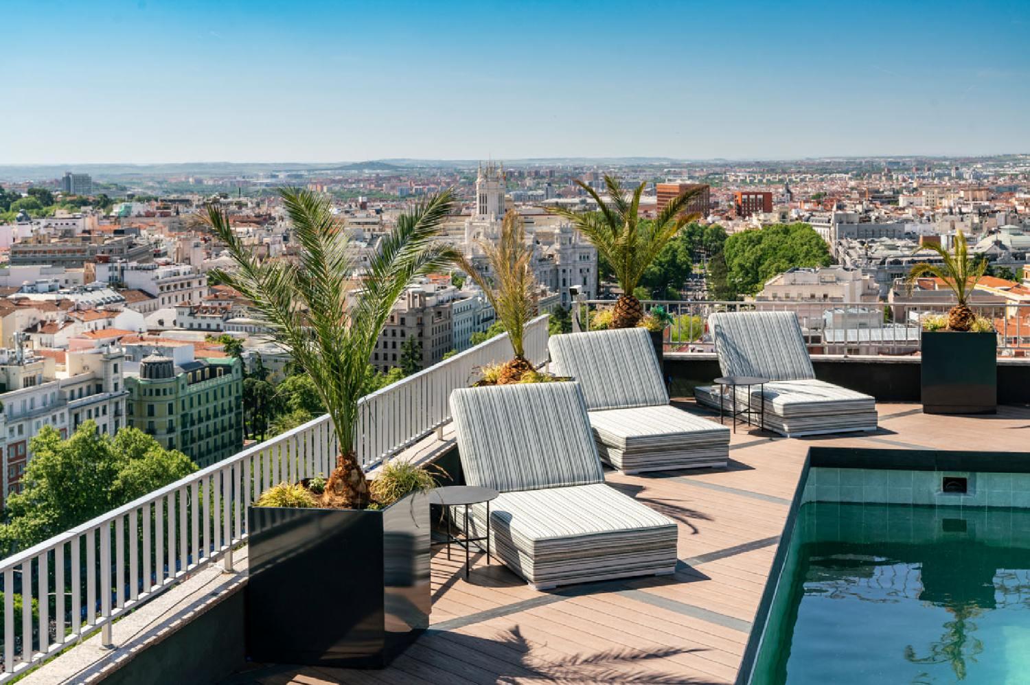 Zona de piscina en la azotea con tumbonas a rayas y palmeras en macetas, con vistas a un extenso paisaje urbano bajo un cielo azul claro, que transmite relajación y lujo.