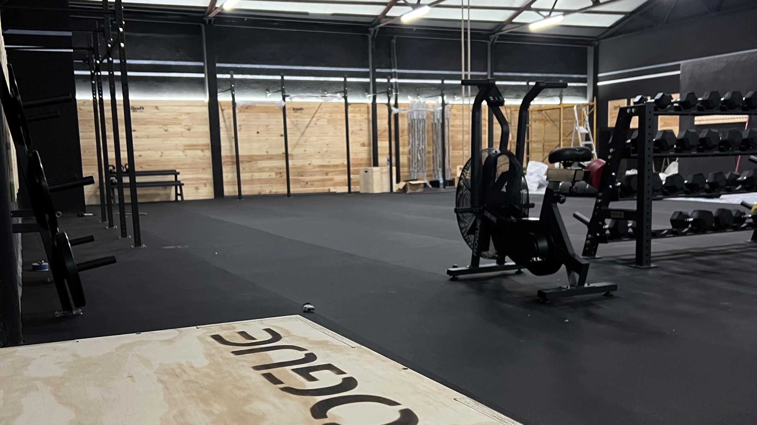 Un amplio gimnasio con suelo de goma cuenta con una máquina de remo y una estantería con mancuernas. Las paredes de madera y los techos altos crean un ambiente moderno y diáfano.