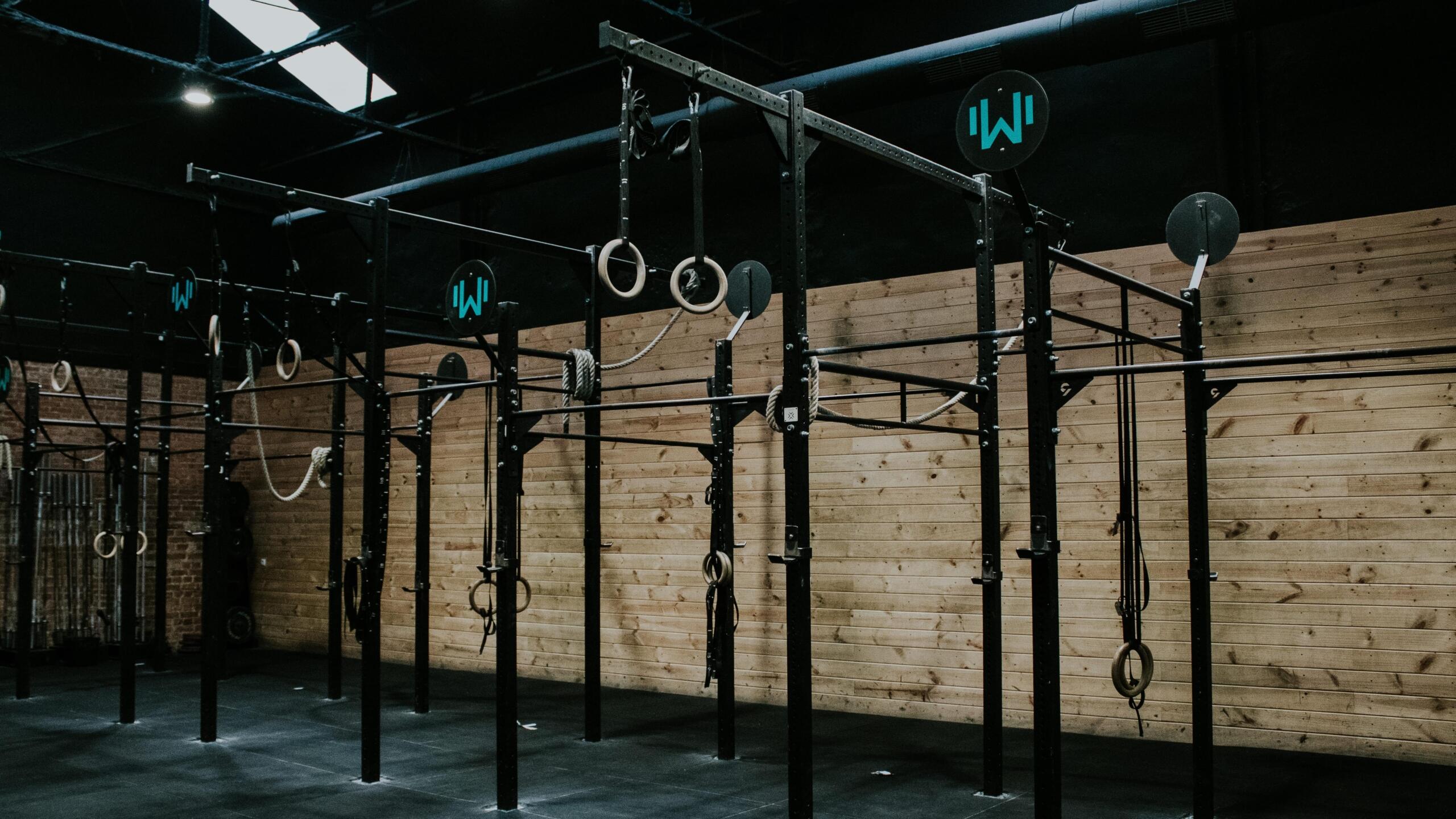Un gimnasio con poca luz cuenta con estructuras de entrenamiento cruzado de metal negro, con anillas de gimnasia y pesas. Las paredes de madera le dan un aire rústico y robusto.