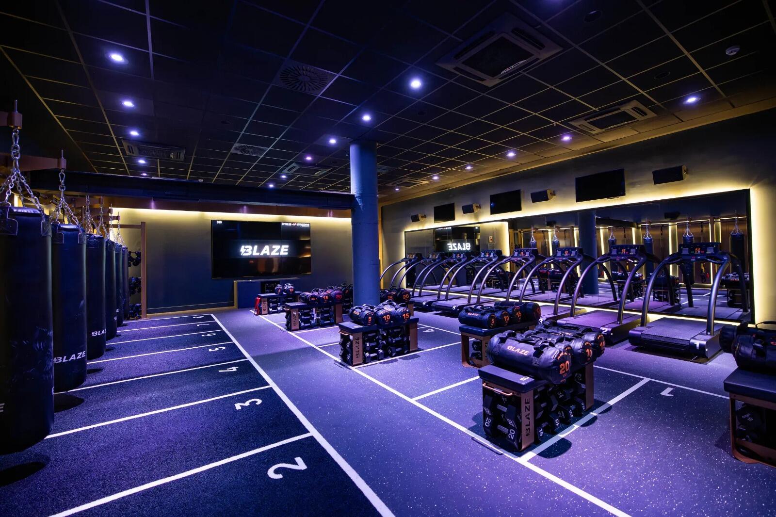 Un gimnasio moderno, iluminado con luces de neón moradas, cuenta con sacos de boxeo alineados, cintas de correr y bancos de pesas. El ambiente es enérgico y elegante.