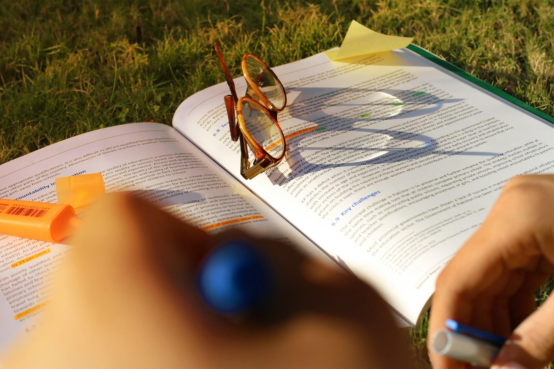 Libro abierto sobre la hierba, con gafas, texto subrayado y notas adhesivas. Una persona escribe con un bolígrafo azul, lo que evoca un ambiente de estudio al aire libre.