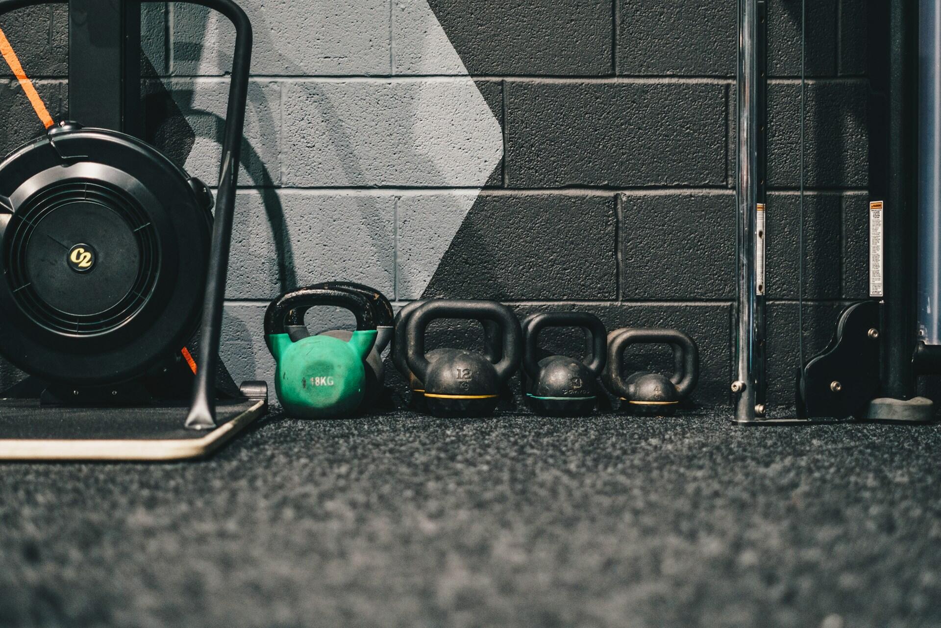 Varias pesas rusas de distintos tamaños, entre ellas una llamativa de color verde con la inscripción «18 kg», están cuidadosamente alineadas en el suelo del gimnasio, junto a una pared de ladrillo gris.