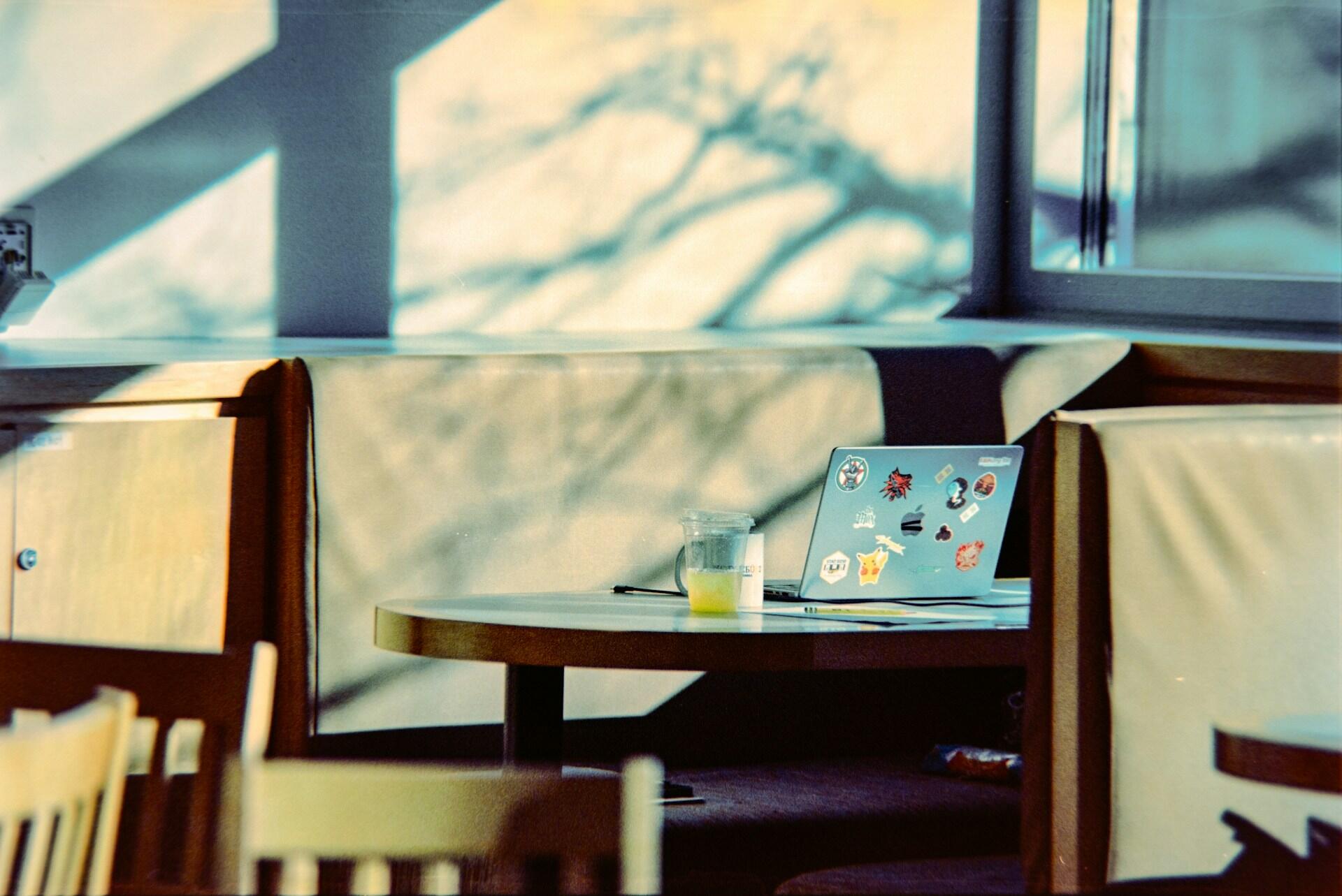 Una acogedora mesa de cafetería bañada por la cálida luz del sol, con un portátil decorado con pegatinas y un vaso de zumo de naranja medio lleno sobre la mesa, lo que crea un ambiente relajado y acogedor.