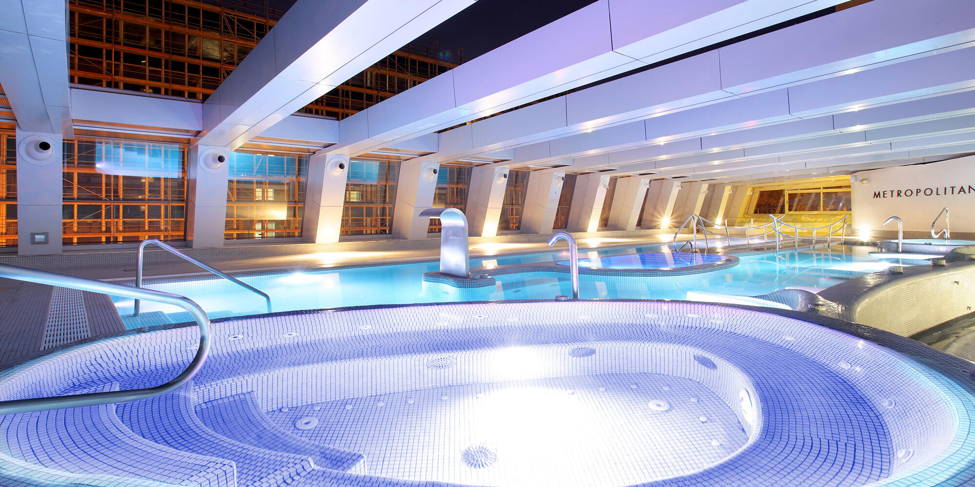 Amplia zona de piscina cubierta de diseño moderno, con un jacuzzi circular iluminado y una piscina rectangular. Ambiente relajante con una suave iluminación azul.
