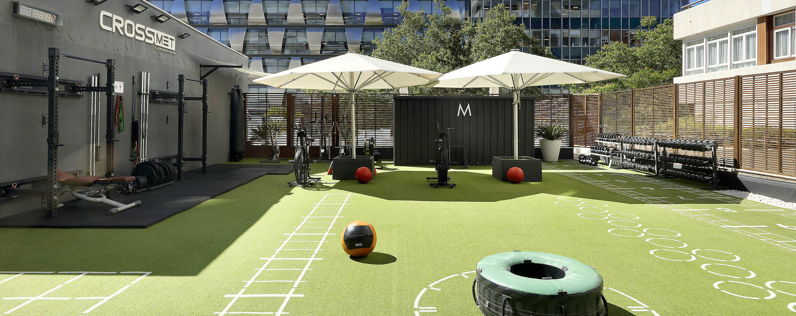 Gimnasio al aire libre de CrossFit con equipamiento como pesas y balones medicinales sobre césped artificial. Dos grandes sombrillas y edificios modernos crean un espacio urbano para hacer ejercicio.