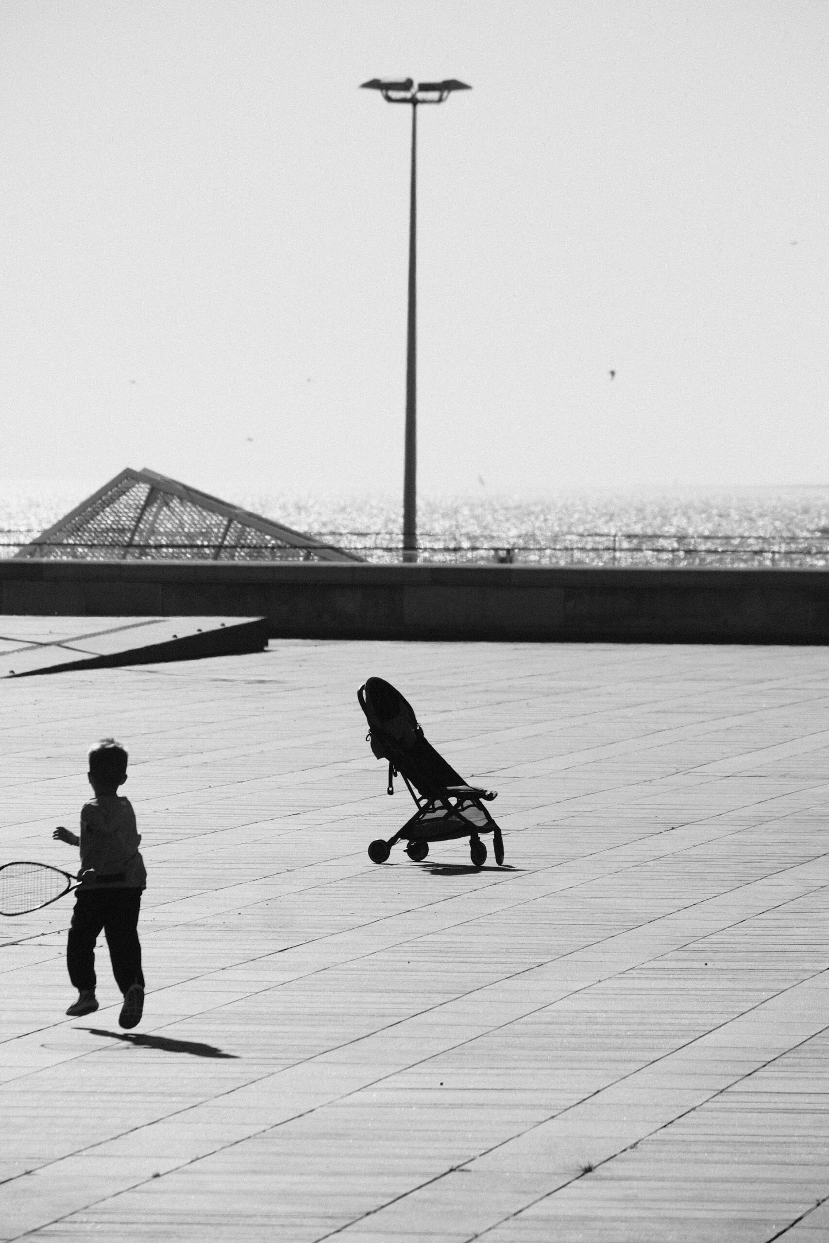 Imagen en blanco y negro de un niño corriendo con una raqueta junto a un cochecito vacío en un paseo marítimo bañado por el sol, con el mar brillando al fondo.