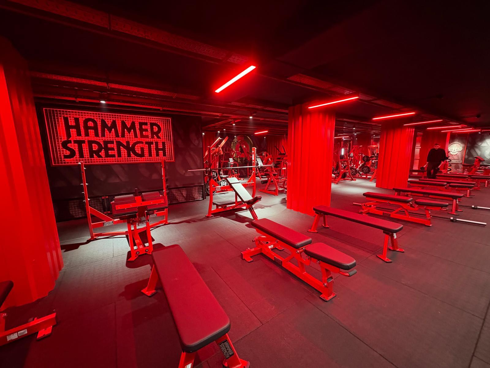 Una sala de gimnasio bañada en luz roja, con varios bancos y máquinas de musculación. Un cartel de «Hammer Strength» ocupa un lugar destacado, lo que crea un ambiente intenso.