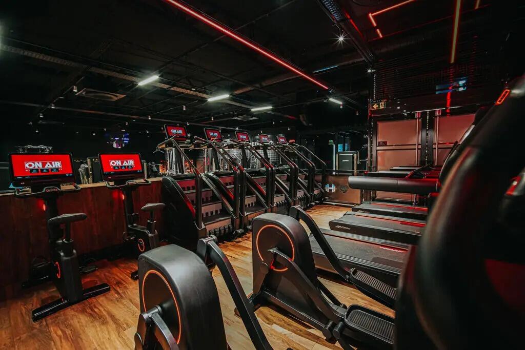 Gimnasio moderno con iluminación tenue, con filas de cintas de correr y bicicletas estáticas. Las líneas de color rojo vivo y las pantallas con el letrero «On Air» le dan un aire enérgico y de alta tecnología.