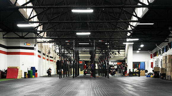 Interior de un gimnasio espacioso de estilo industrial, con techos altos, iluminación tenue, estanterías negras para el equipamiento y gente realizando ejercicios físicos. Ambiente elegante y moderno.