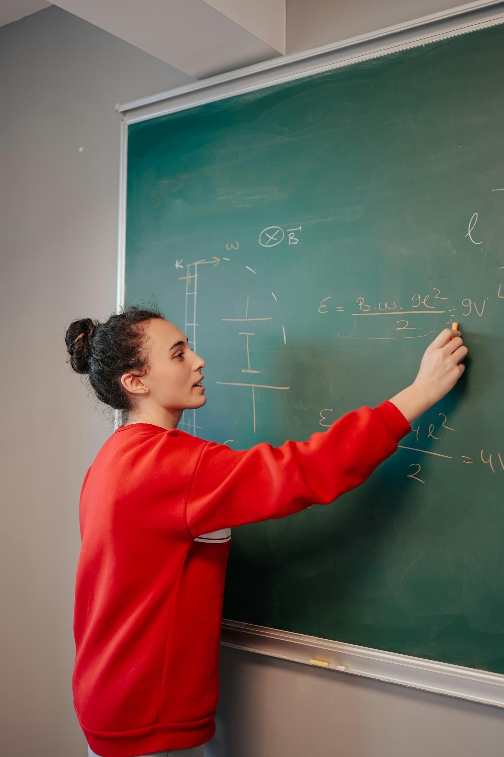 Una persona con una sudadera roja escribe fórmulas en una pizarra verde, demostrando un concepto de física en un aula.