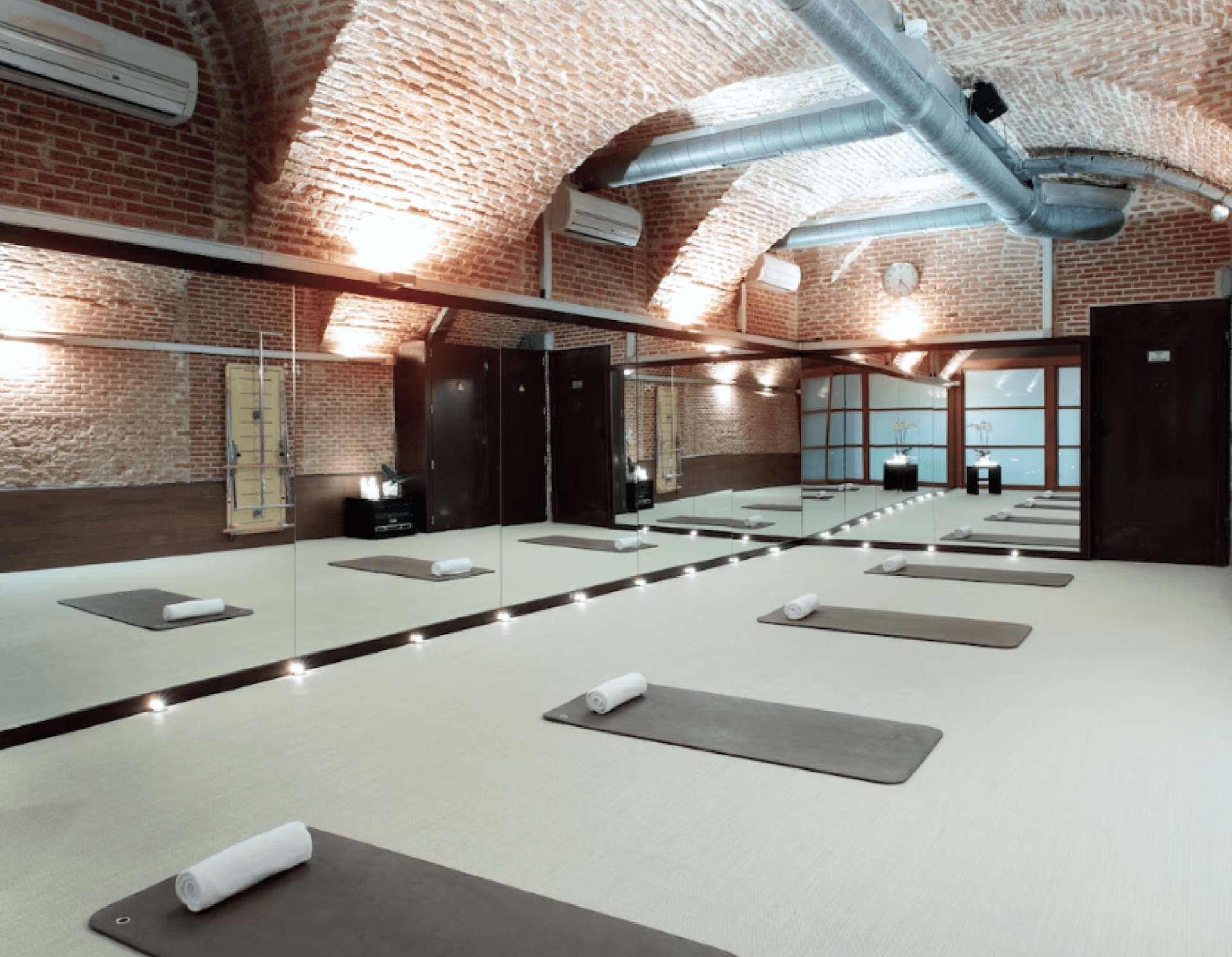 Estudio de yoga con techos abovedados de ladrillo y paredes acristaladas. Las colchonetas, con toallas enrolladas, están distribuidas uniformemente por el suelo, creando un ambiente tranquilo.