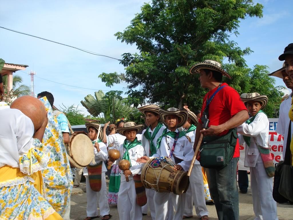 Una colorida escena callejera en la que aparecen niños con trajes tradicionales, tocando instrumentos y preparándose para un evento festivo.