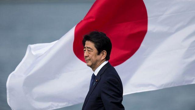 Shinzo Abe vestido con traje posa solemnemente junto a una gran bandera japonesa ondeando al viento. El océano se ve al fondo, lo que transmite un ambiente reflexivo.