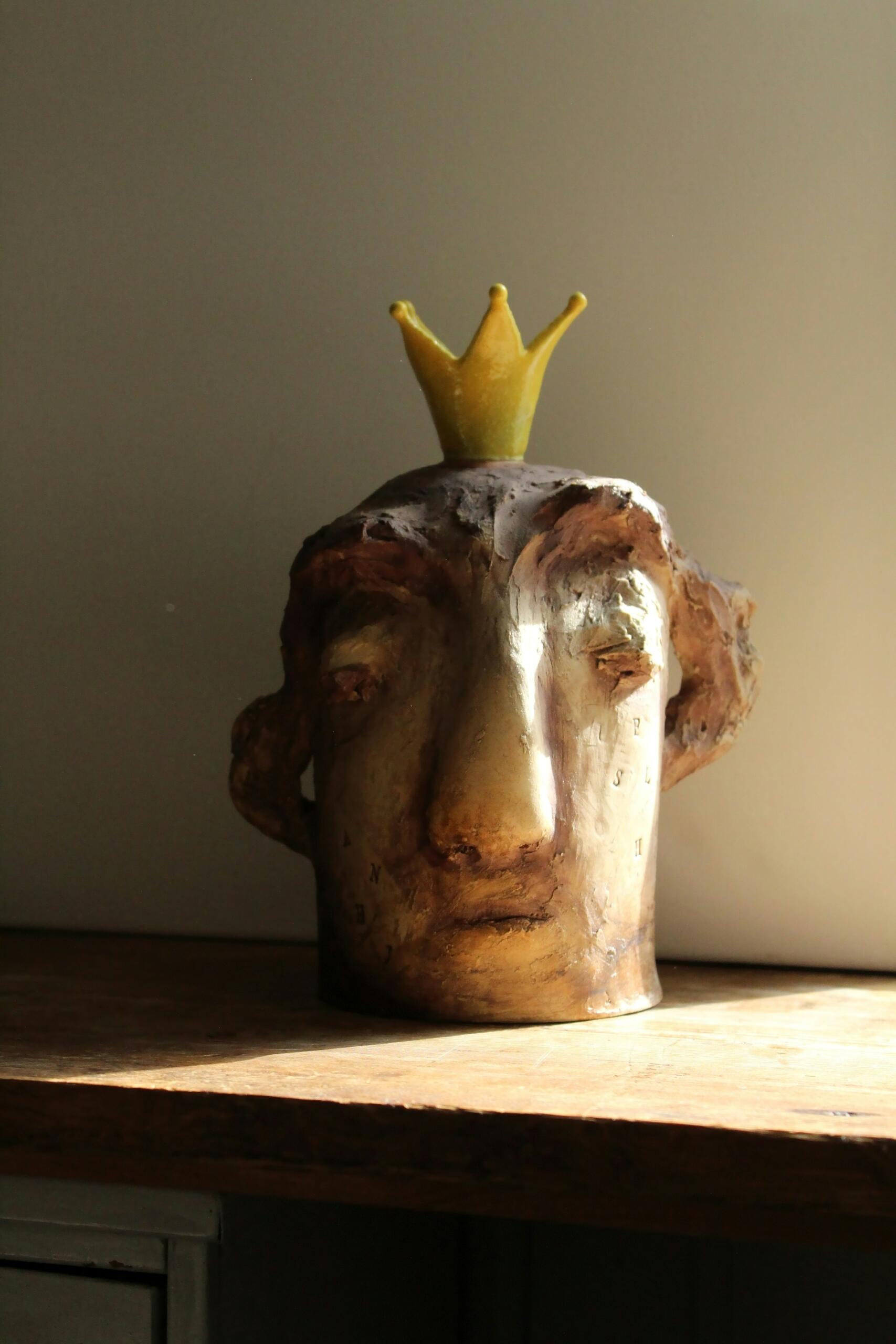 Una cabeza esculpida con expresión serena lleva una corona amarilla, colocada sobre una superficie de madera. Una luz suave proyecta sombras, creando un ambiente contemplativo.