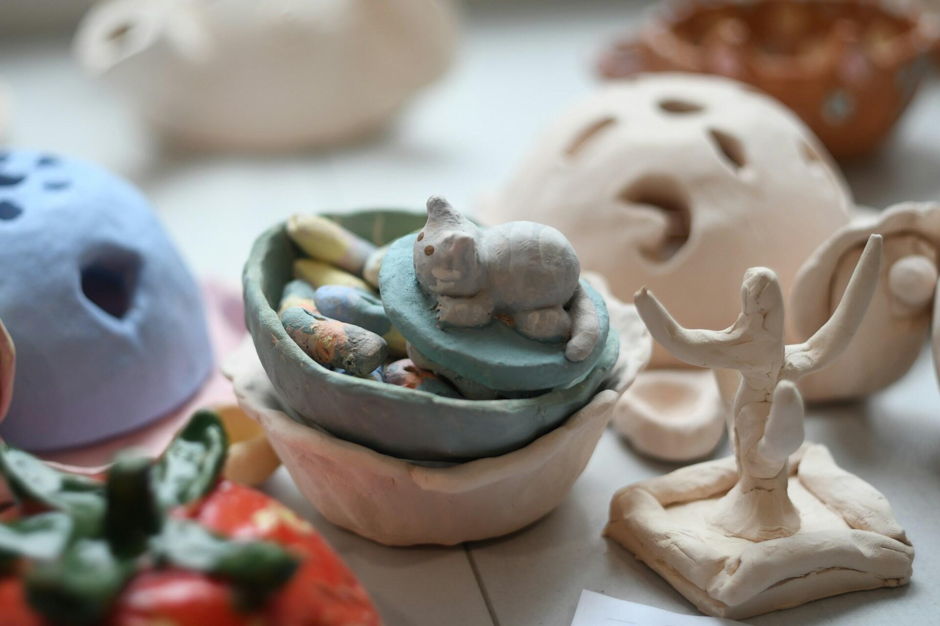 Una colección de pequeñas y coloridas esculturas de arcilla en exhibición. Incluye un gato sobre un cuenco, una figura con los brazos levantados y un objeto rojo parecido a un tomate. Brillante y divertido.