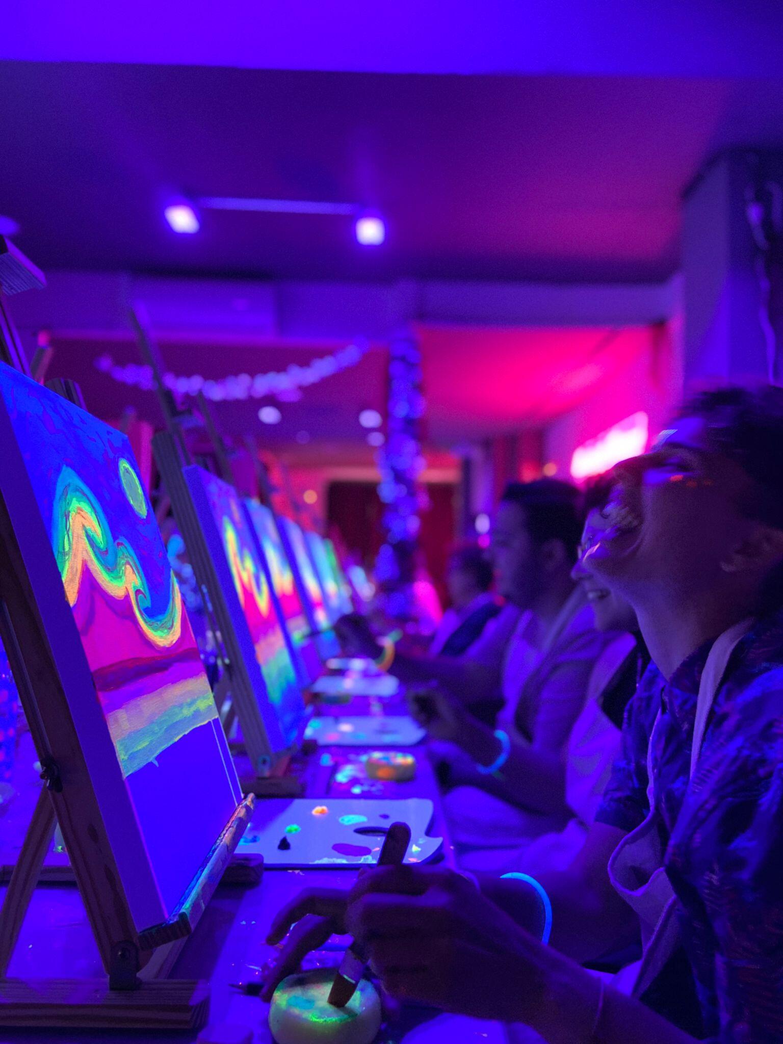 Personas pintando bajo luces de neón en una habitación oscura. Los lienzos brillan con colores vibrantes. Los participantes llevan delantales y pulseras. El ambiente es alegre y creativo.