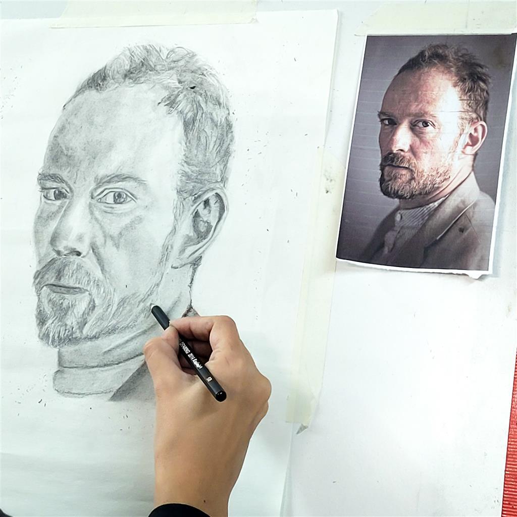 Artista dibujando un retrato realista a lápiz de un hombre barbudo sobre lienzo; junto a él hay una foto de referencia, lo que demuestra su concentración y su talento artístico.