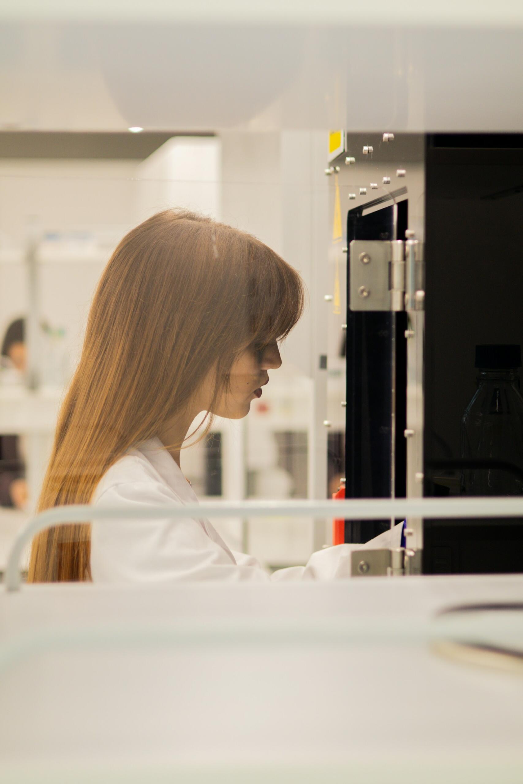 Una mujer con bata de laboratorio maneja un equipo en un laboratorio muy iluminado. Su expresión serena denota concentración y precisión en su trabajo científico.