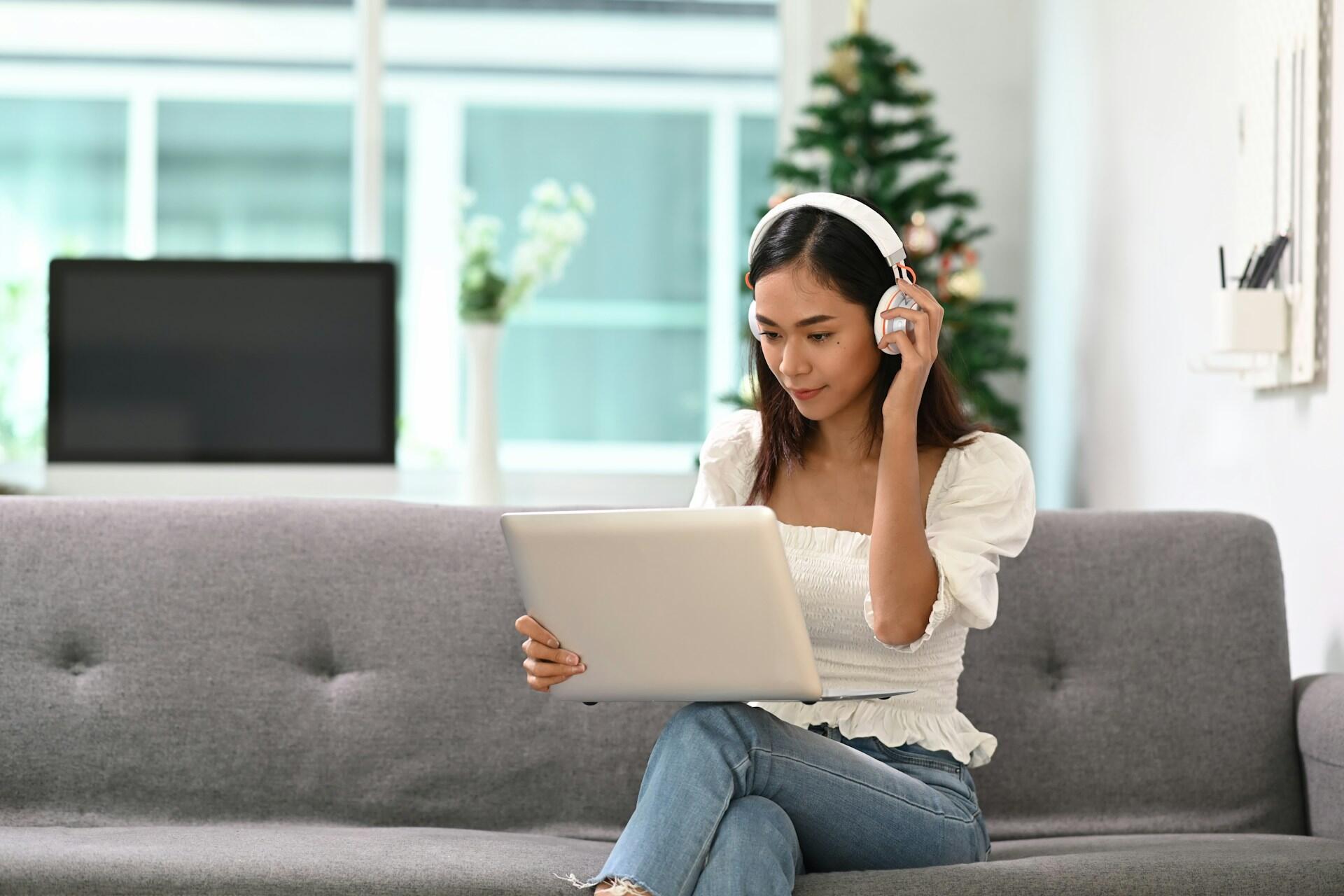 Una persona se sienta en un sofá gris con auriculares, trabajando en una computadora portátil con un árbol de Navidad festivo de fondo.