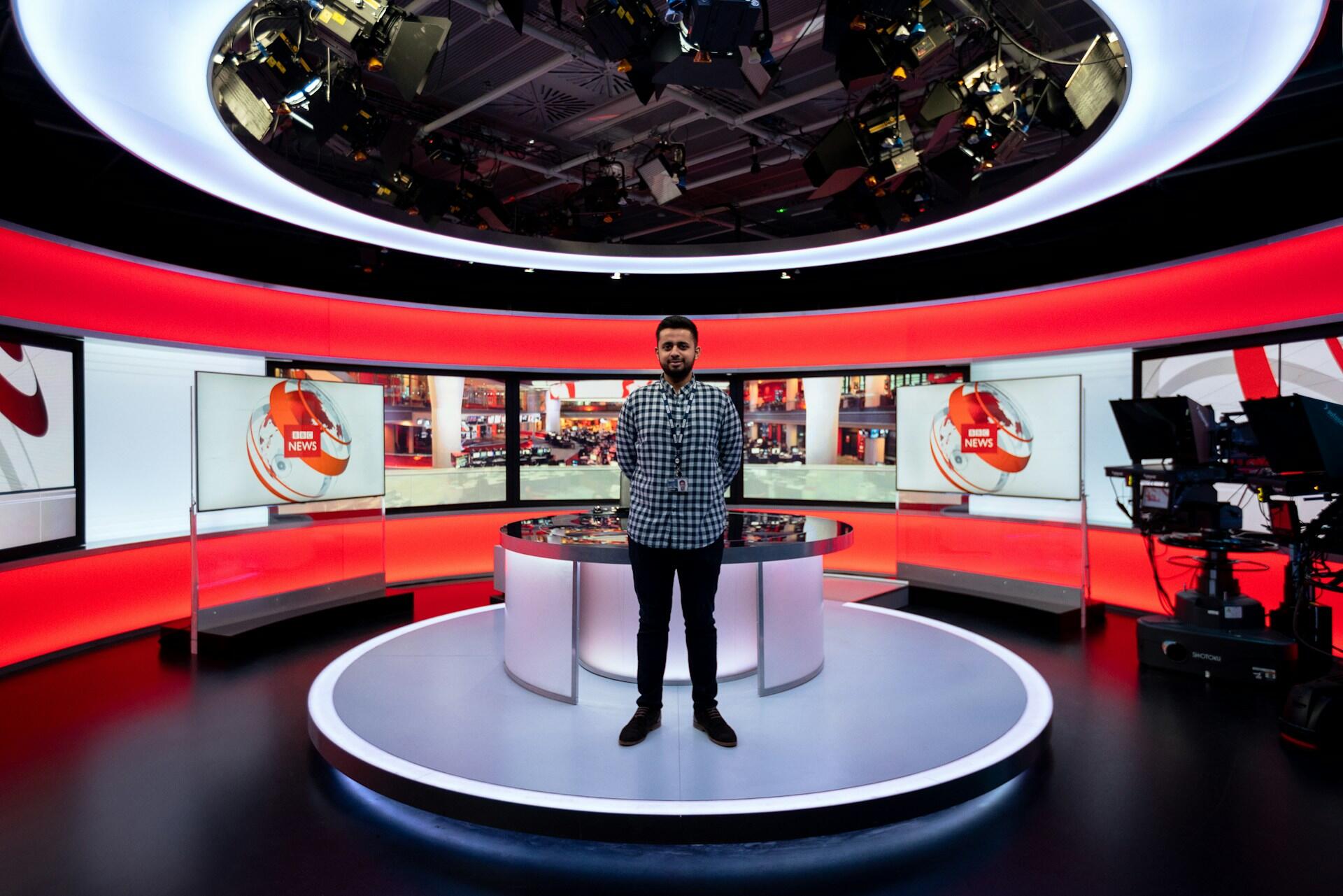Un moderno estudio de noticias con iluminación roja brillante, que cuenta con un escritorio circular, pantallas que muestran gráficos de noticias y cámaras en el costado.