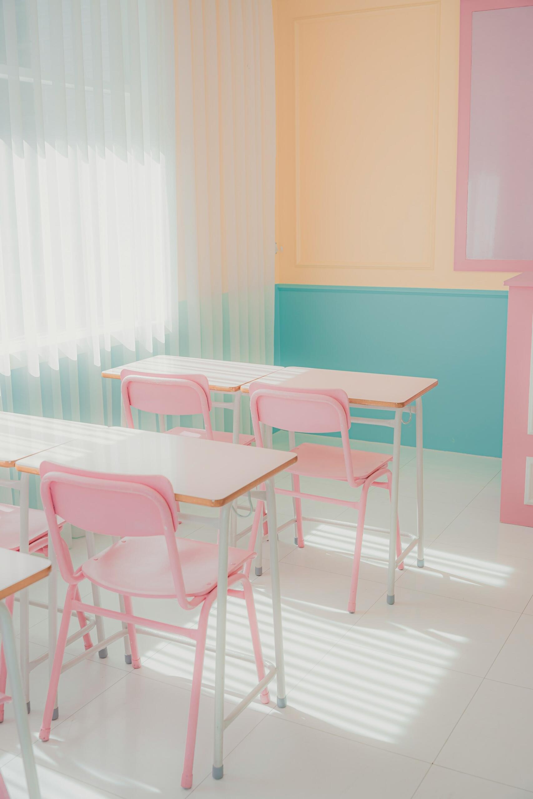 Aula luminosa con colores pastel; pupitres y sillas rosas bajo una luz suave y natural. Las paredes son una mezcla de melocotón, verde azulado y rosa, lo que crea un ambiente alegre y acogedor.