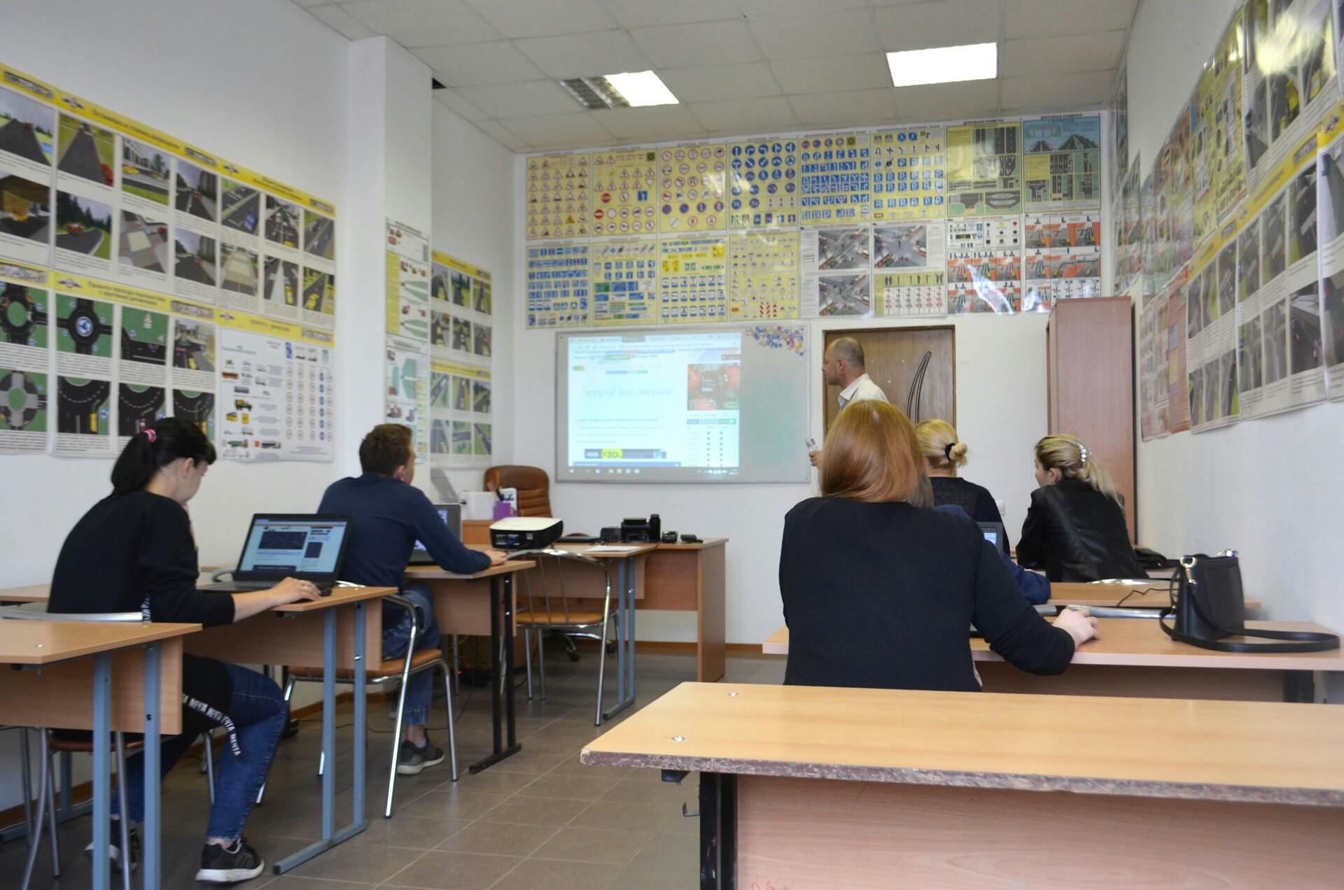 Un aula con cinco personas sentadas en sus pupitres, frente a una pantalla en la que se proyecta una presentación. Las paredes están cubiertas de señales de tráfico y diagramas educativos.