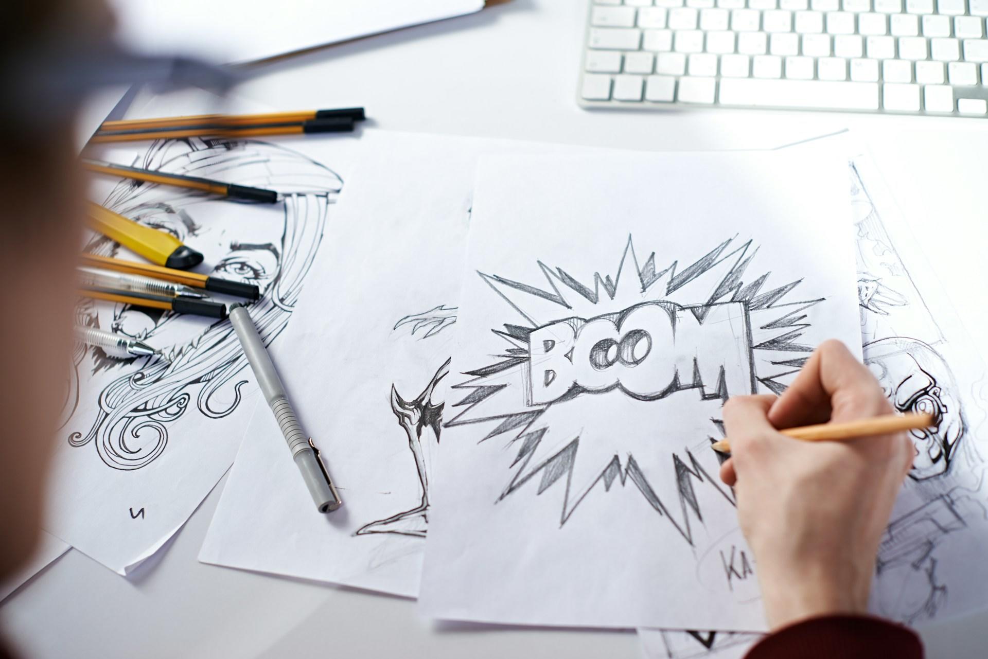 Persona dibujando una explosión de cómic con la palabra «BOOM» en un papel, entre lápices y un teclado.