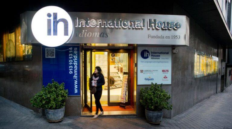 Una mujer sale de la escuela de idiomas International House, iluminada por unas cálidas luces. En la entrada hay un gran cartel con logotipos educativos, flanqueado por macetas con plantas.