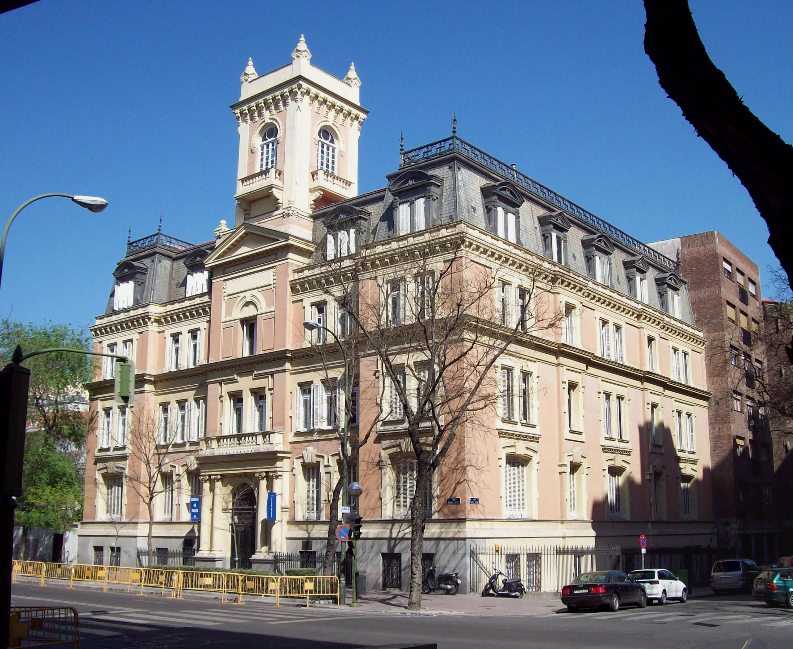 Elegante edificio histórico de color beige con una alta torre central, detalles intrincados y tejados abuhardillados, recortado contra un cielo azul claro y árboles desnudos.
